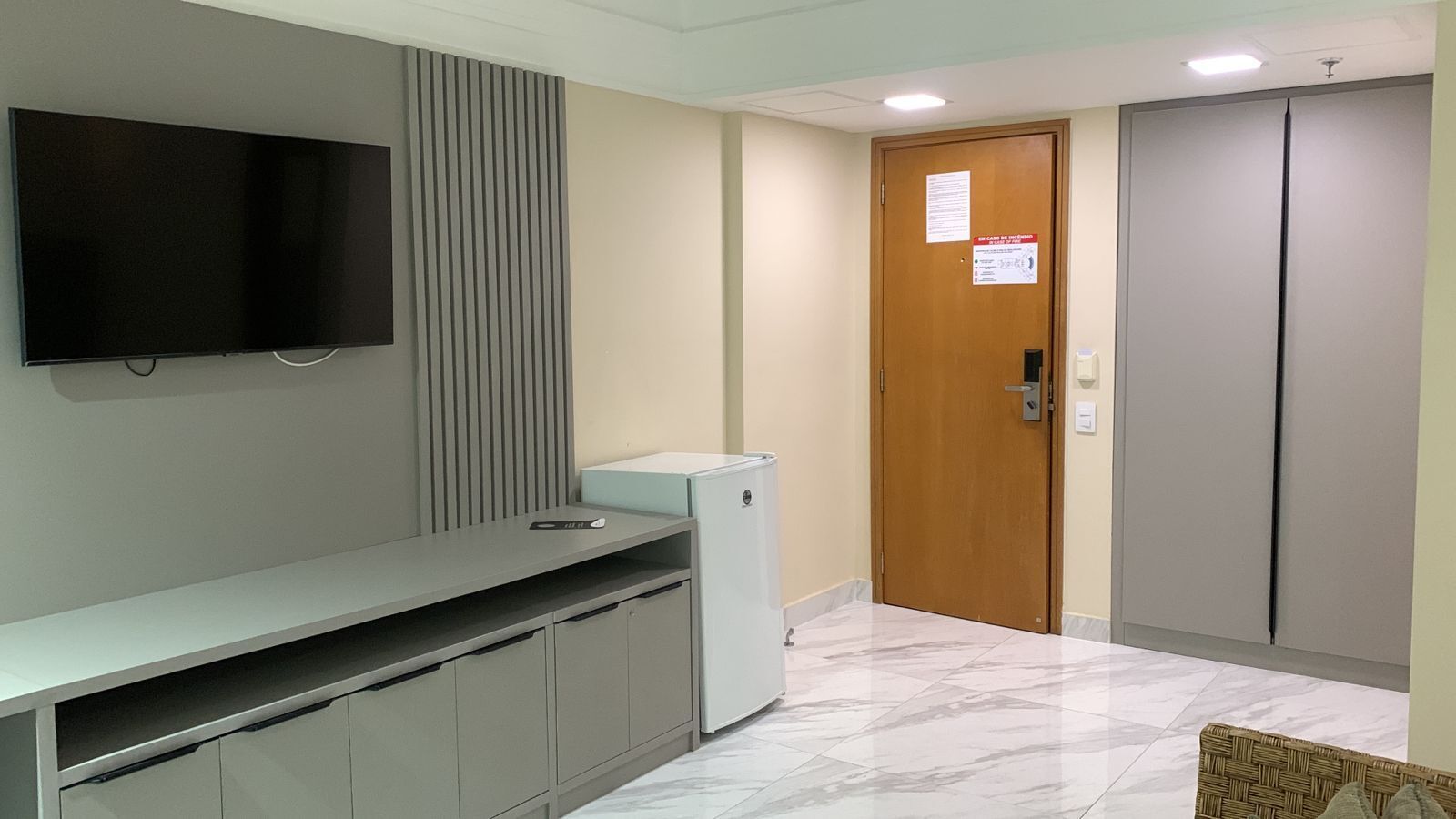 A hotel room with a flat-screen TV mounted on the wall, a small refrigerator, a wooden door with a sign, and a wardrobe.