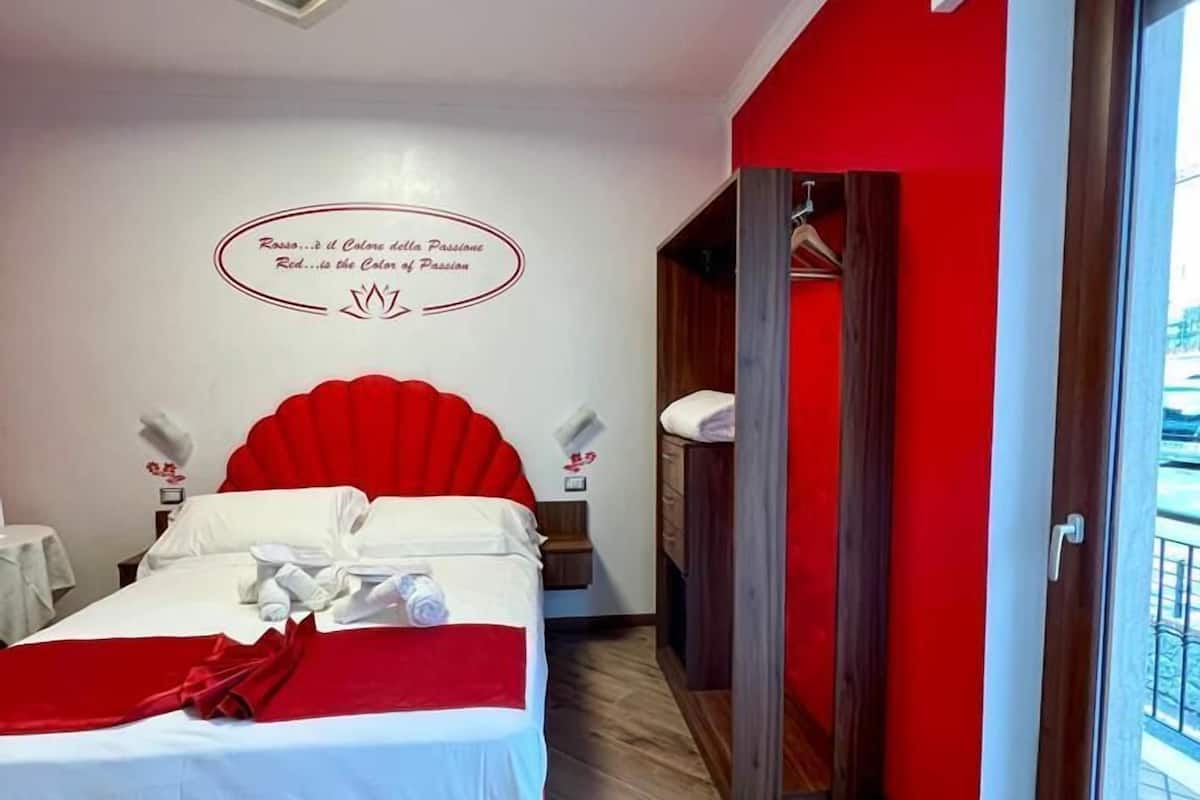 A hotel room with a bed, a red accent wall, a ceiling light, and a view through a window.