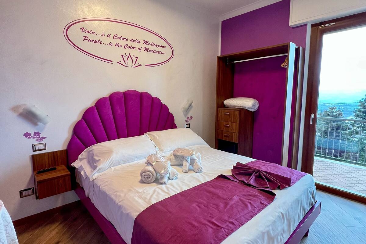 A bedroom with a purple bed, white pillows, and a ceiling light. There is a wall with a quote and a small shelf with a lamp.
