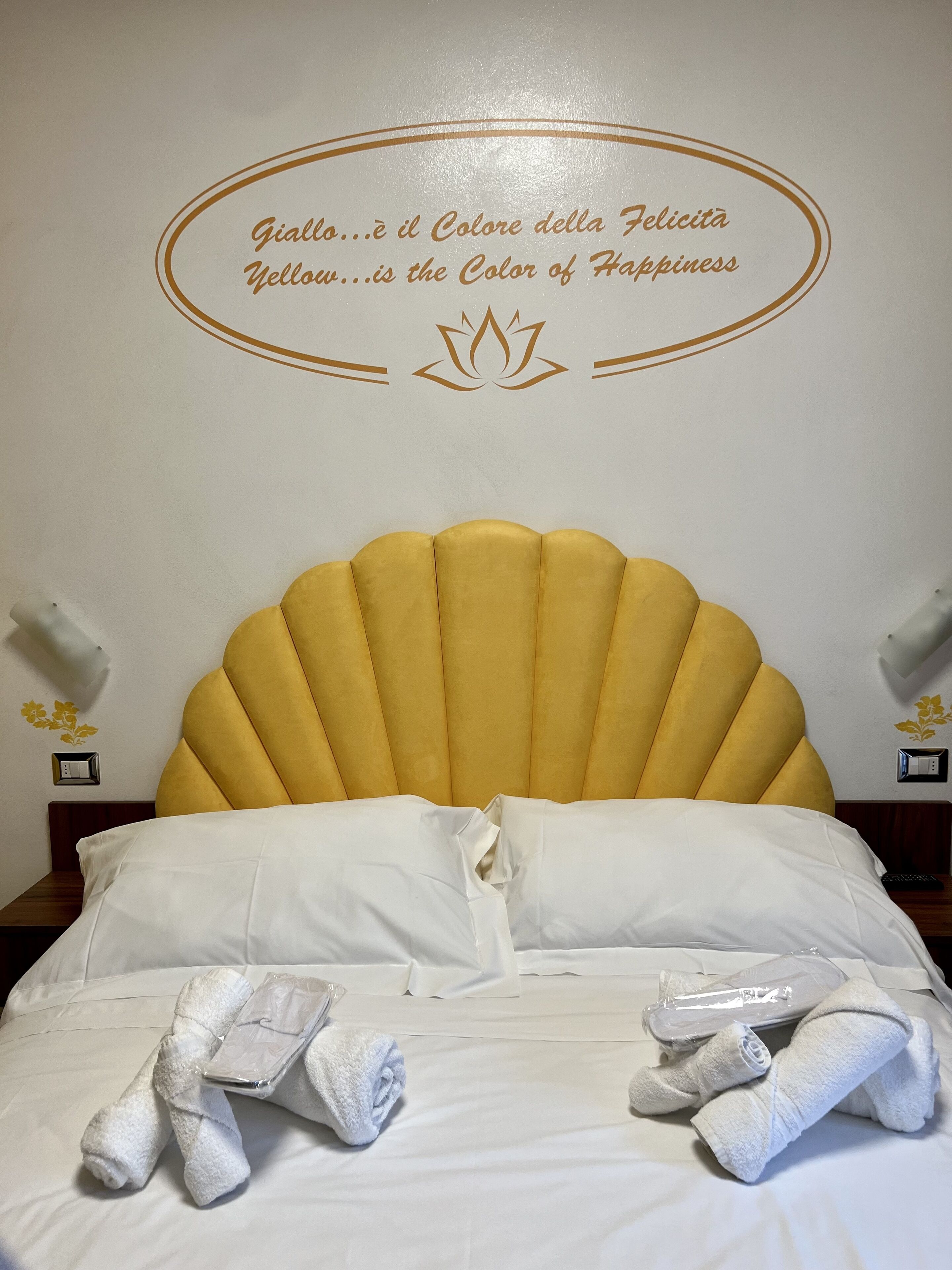 A bed with a yellow headboard, white pillows, and rolled towels.