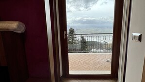 Una habitación de hotel con una cama, una toalla morada y vistas a un balcón con barandilla y un cielo nublado.
