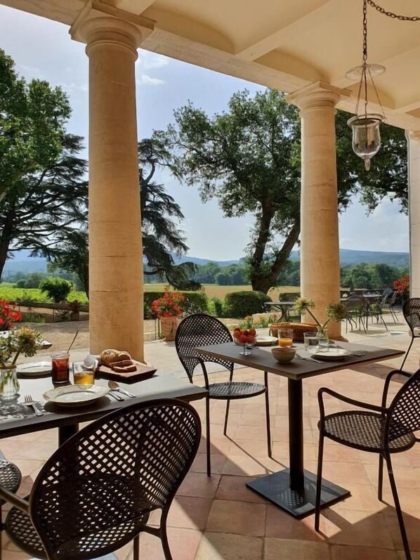 Un área de comedor al aire libre con una mesa lista para dos personas, rodeada de columnas y con vistas a un paisaje pintoresco.