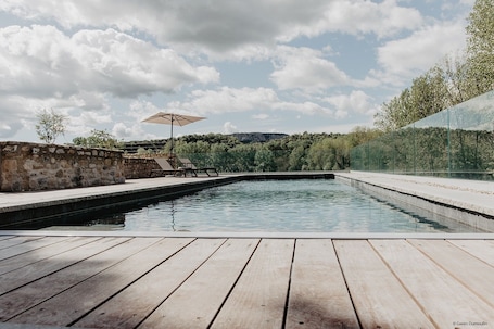 A swimming pool with wooden decking, a stone wall, and a glass fence.