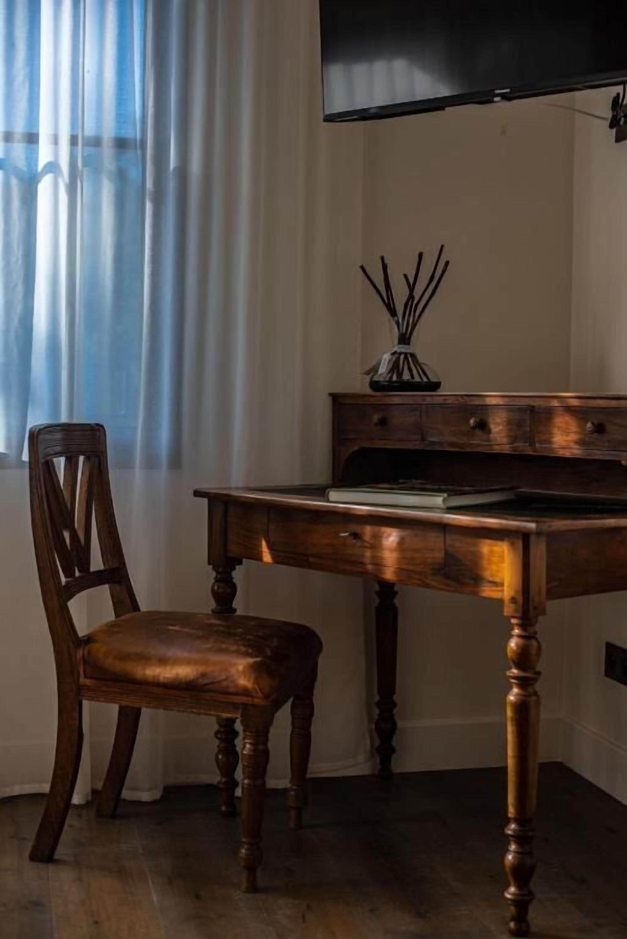 A wooden desk with a television mounted above it, a chair, and a vase with sticks on the desk.