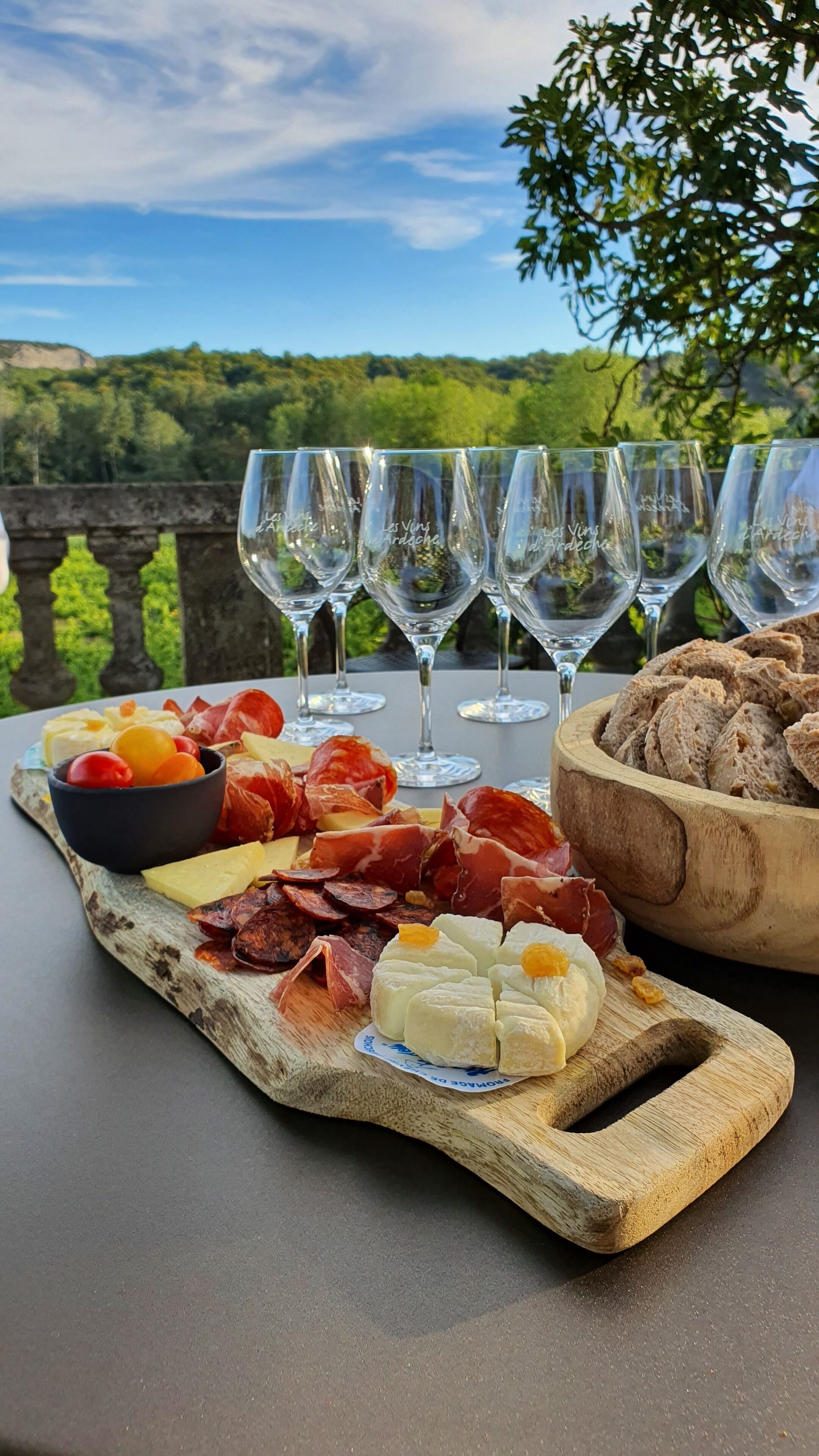 A charcuterie board with various meats and cheeses, accompanied by wine glasses and bread, set outdoors with a scenic view in the background.
