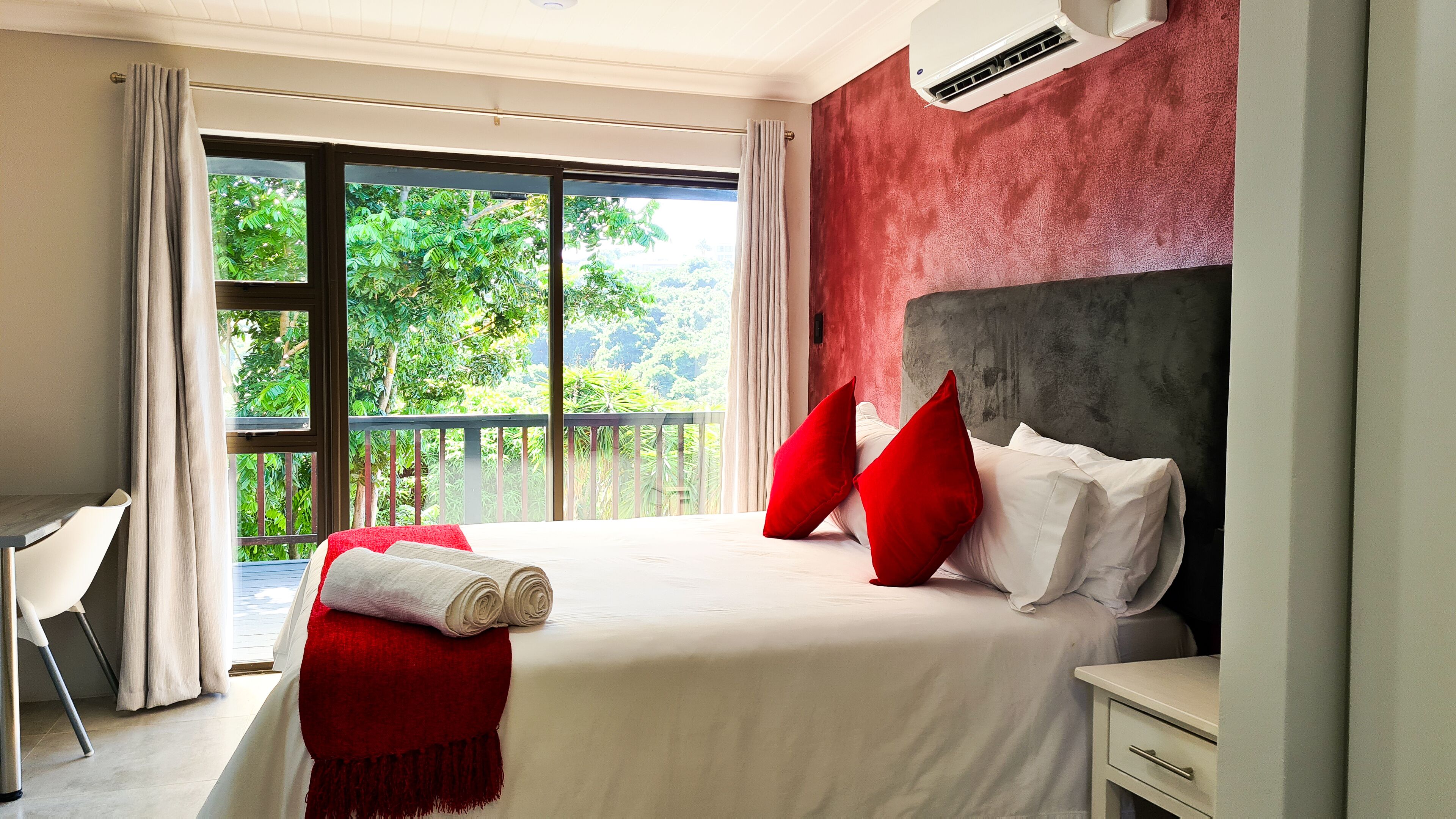 A hotel room with a large bed, a balcony with a view of greenery, a desk, and a modern air conditioning unit.