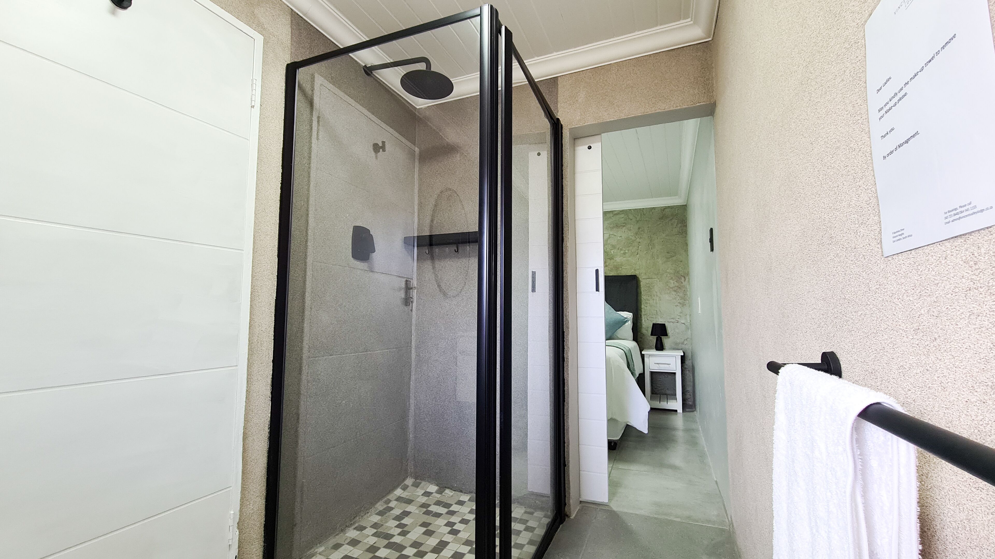 A modern bathroom with a glass-enclosed shower, a white tiled floor, and a visible bed in the adjacent room.