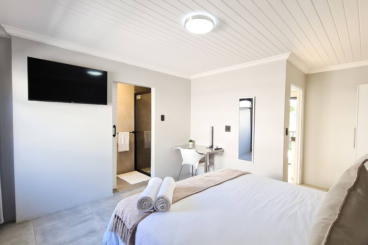 A hotel room with a bed, a desk, a chair, a TV, and a shower area.