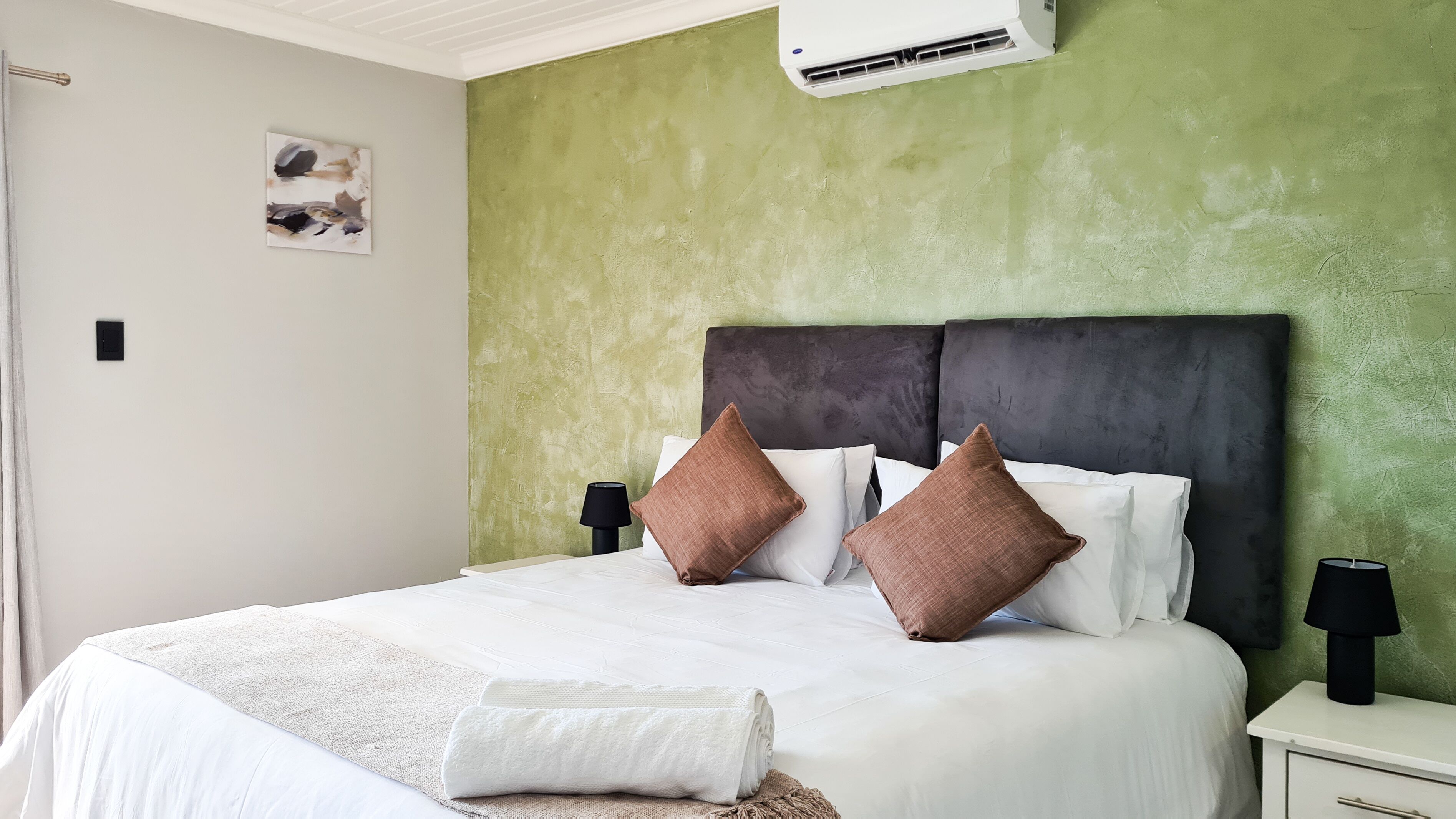 Een opgemaakt bed met witte lakens en twee bruine kussens, een witte nachtkastje met een lamp, en een groene, structuurrijke muur.