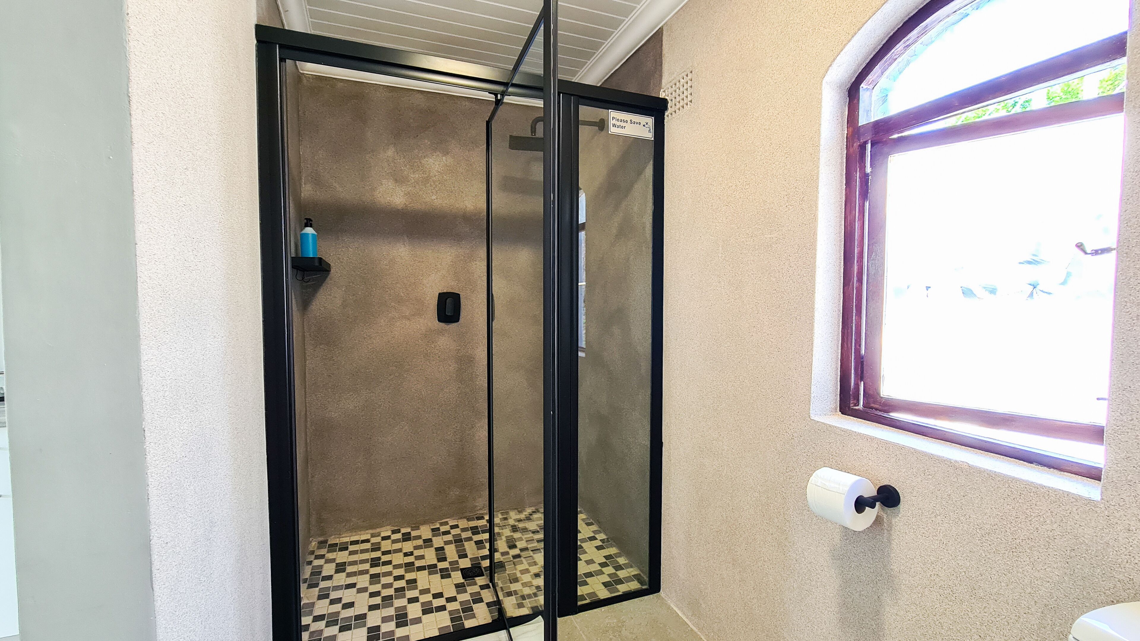 A glass-enclosed shower with a checkered floor, a window with a view, and a sign instructing to save water.