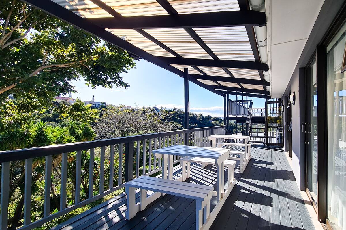 A covered outdoor balcony with wooden furniture and a view of the surrounding greenery and buildings.