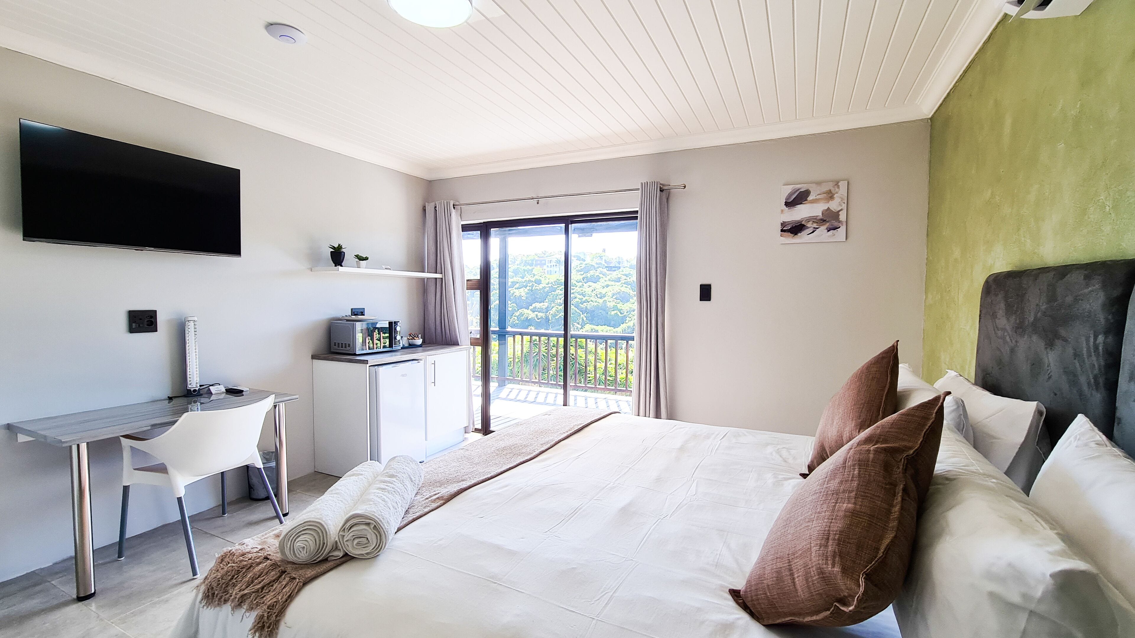 A hotel room with a bed, a desk, a TV, and a balcony with a view of trees.