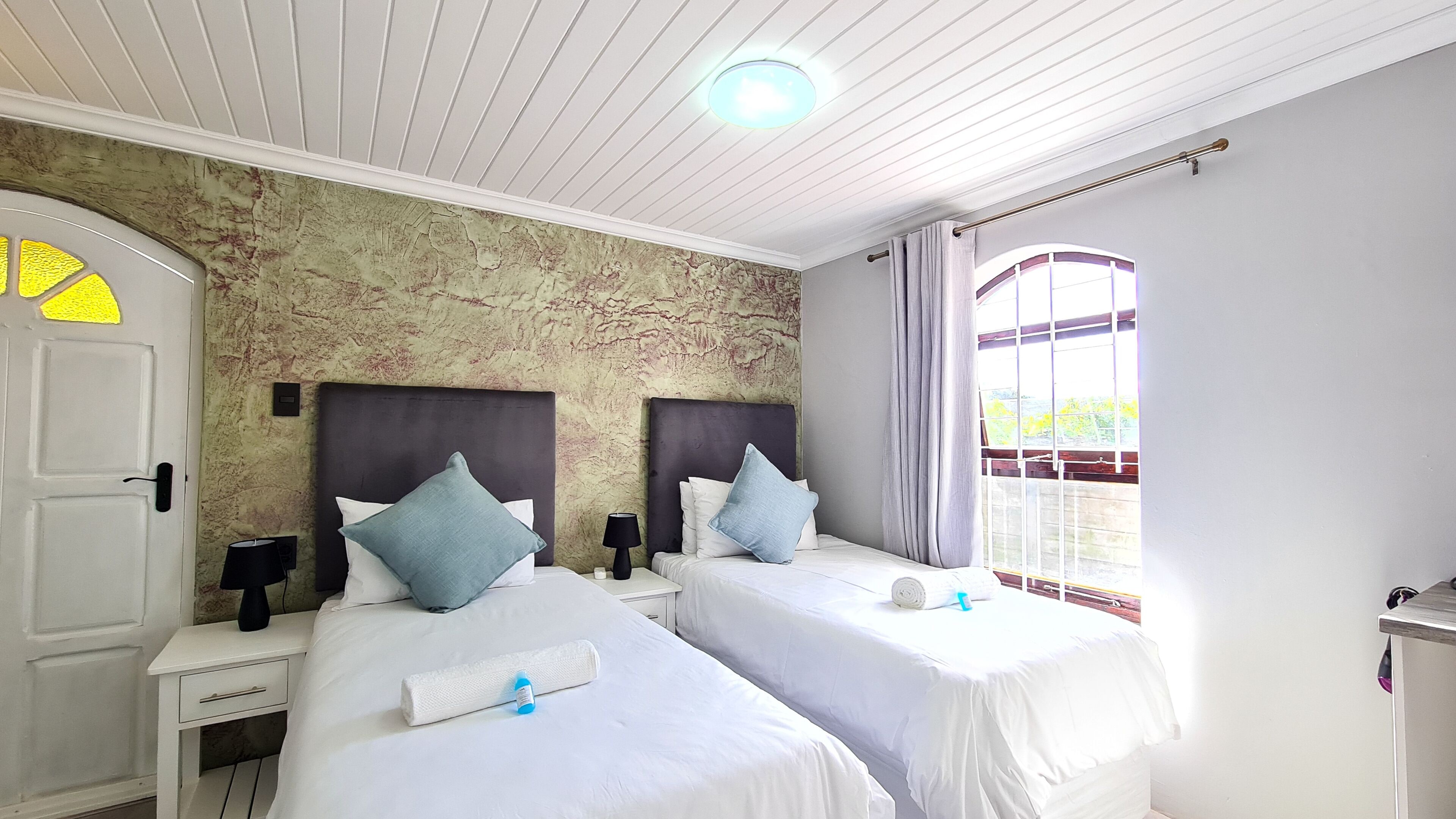 A hotel room with two beds, a white door, a nightstand with a lamp, and a view of greenery through a window.