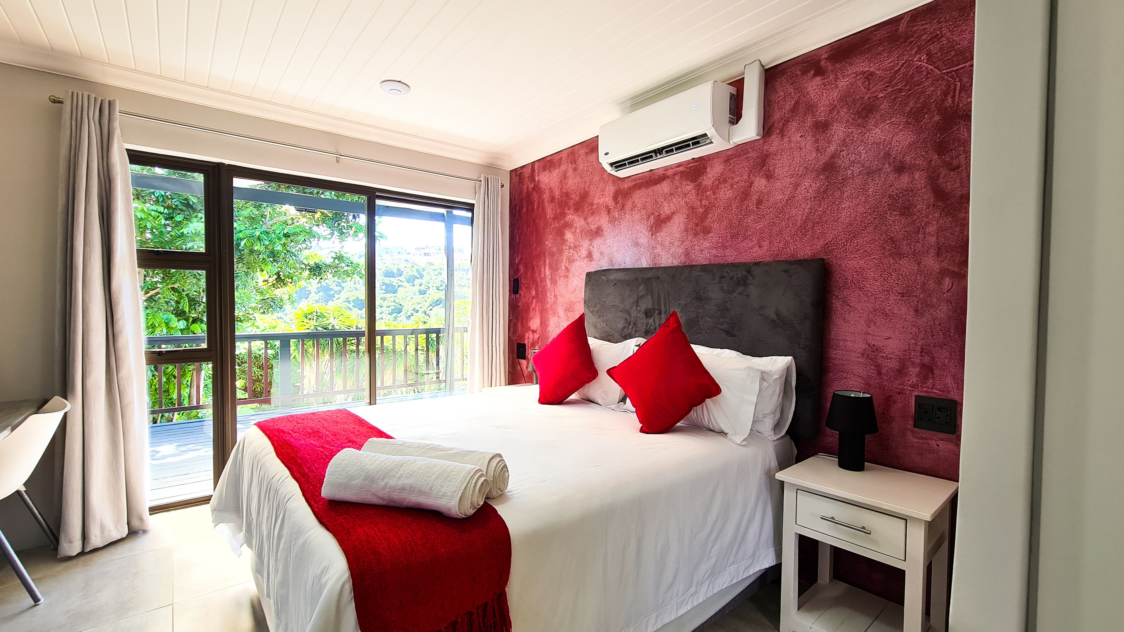 Quarto com uma cama grande, almofadas vermelhas e brancas, um cobertor vermelho, uma mesa de cabeceira, um ar condicionado e vista para o verde através de uma porta de correr.
