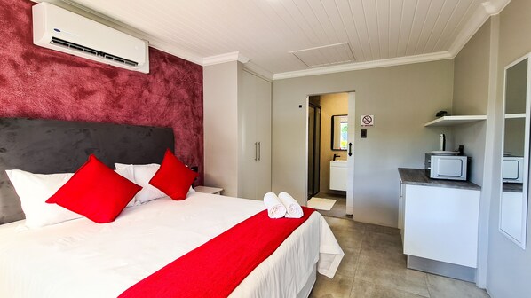 Habitación de hotel con cama, una manta roja, dos almohadas rojas, cabecera blanca, pared roja, armario blanco, microondas y cartel de prohibido fumar.