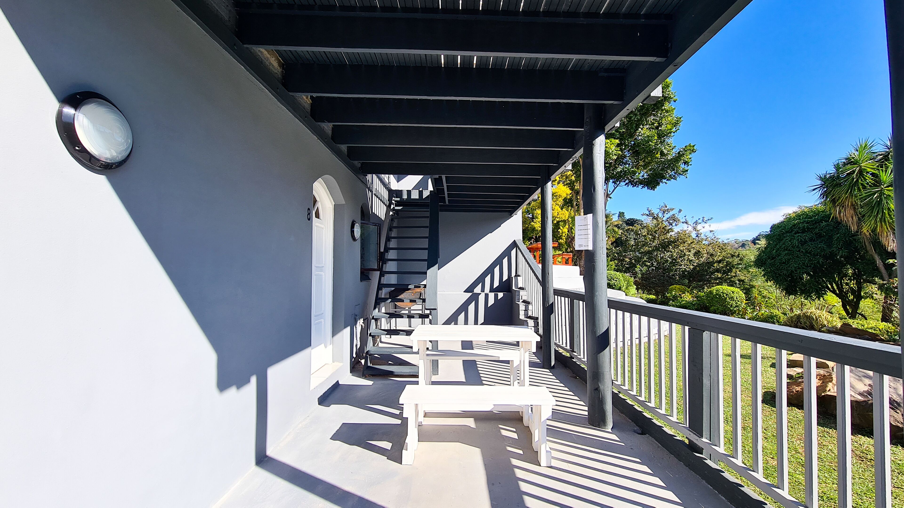 A covered outdoor area with a staircase leading to a second floor, a white bench, and a railing overlooking a garden.