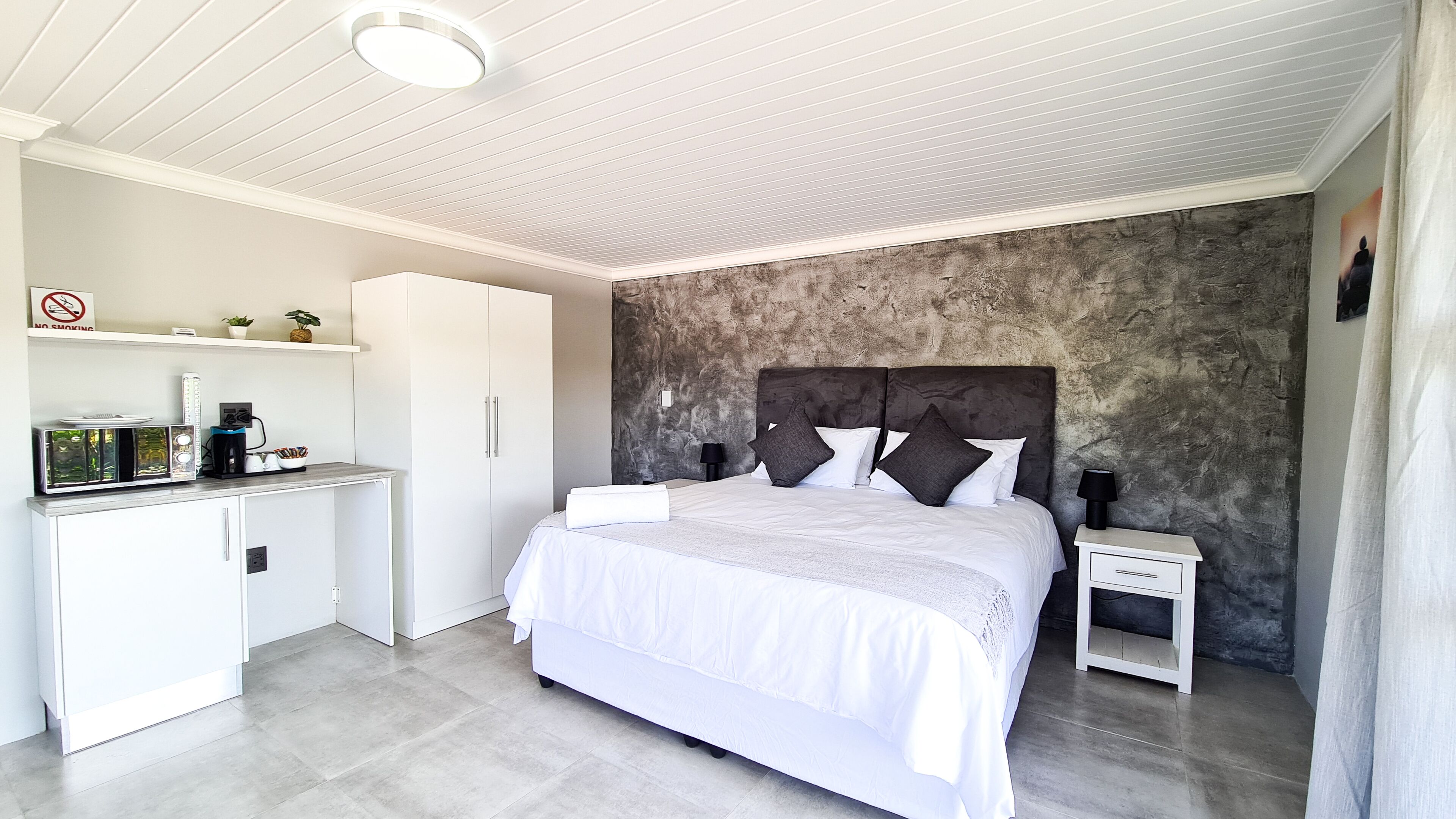 A bedroom with a bed, bedside tables, a wardrobe, a desk with a coffee machine, and a wall with a textured finish.