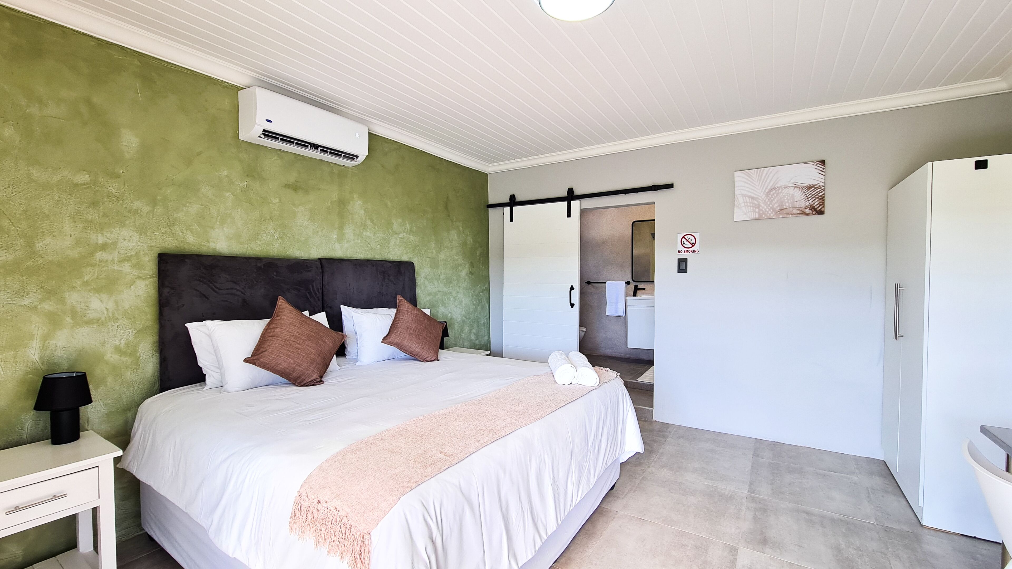Ein Schlafzimmer mit Bett, Kissen, einem Nachttisch, einer Lampe, einer Klimaanlage, einem Handtuch, einer Tür und einem an der Wand befestigten Schild.