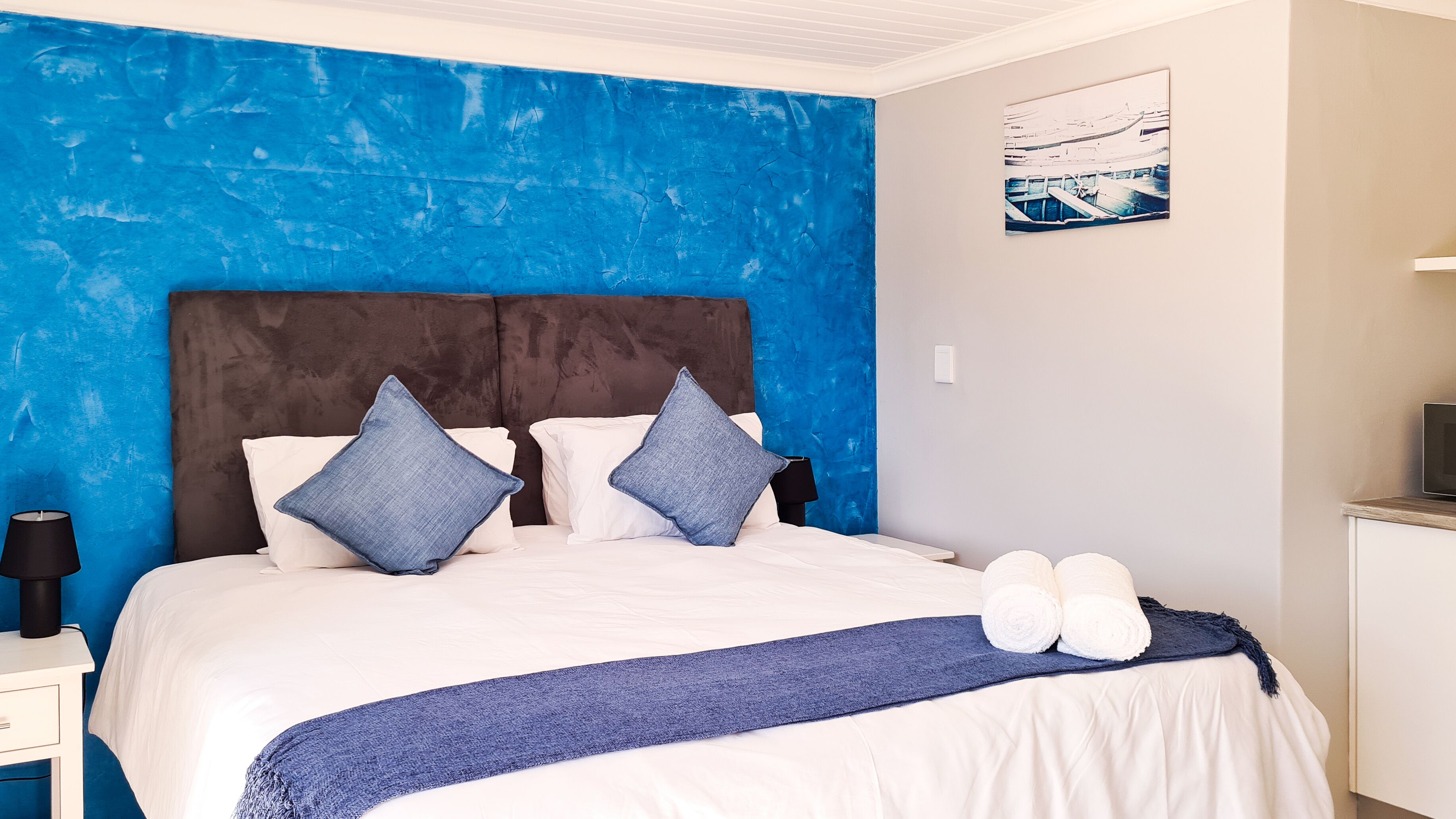 A bedroom with a blue accent wall, a brown headboard, two pillows, a white bedspread, a blue throw blanket, and two rolled towels on the bed.