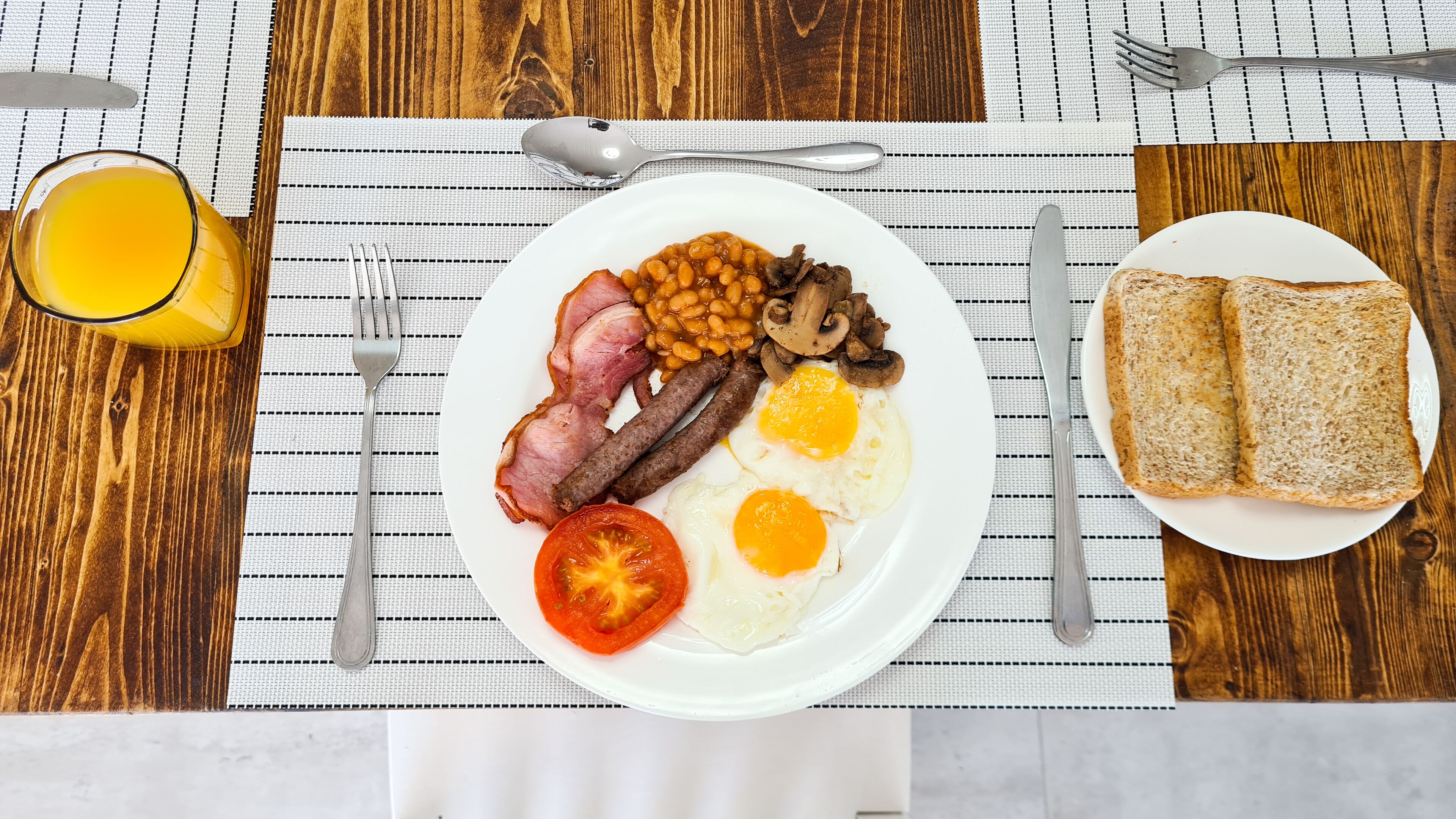 Un piatto per la colazione con uova, salsicce, fagioli, funghi e un pomodoro su un piatto bianco, accompagnato da pane tostato e un bicchiere di succo d'arancia su un tavolo di legno.