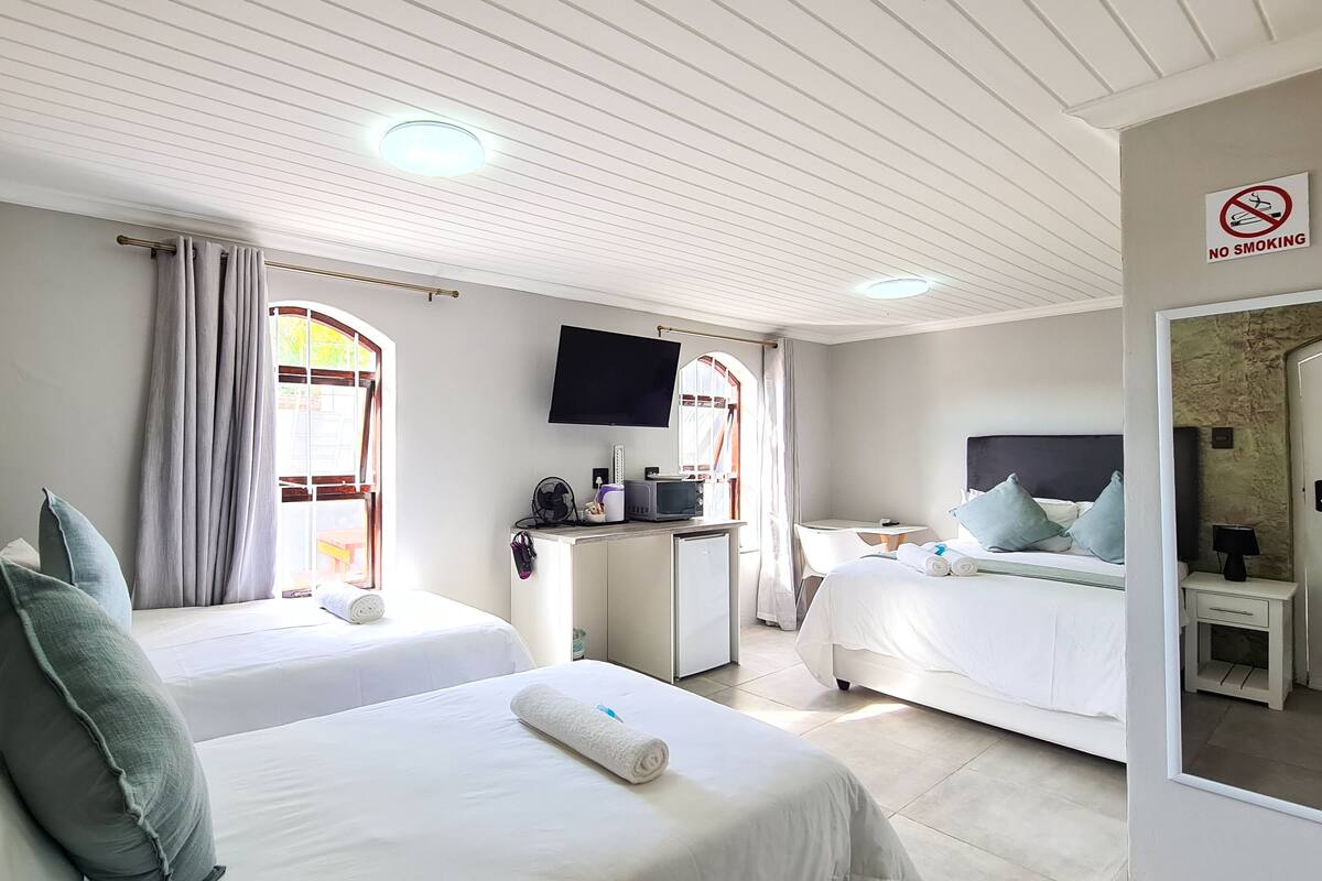 A hotel room with two beds, a desk with a flat-screen TV, a small table, and a no-smoking sign.