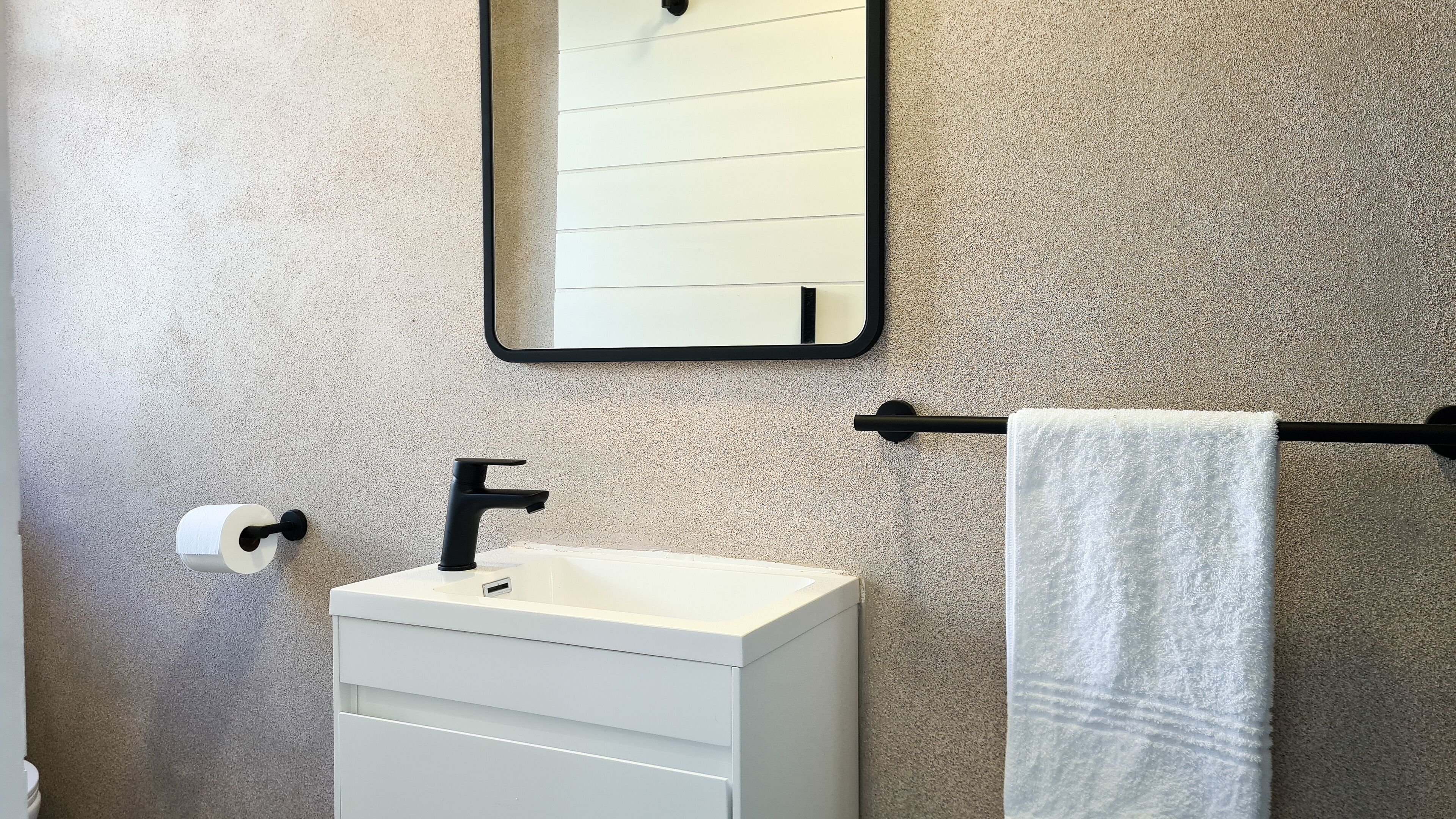 Une salle de bain avec un lavabo blanc, un robinet noir, un porte-serviettes avec une serviette blanche, un porte-rouleau de papier toilette et un miroir avec un cadre noir.