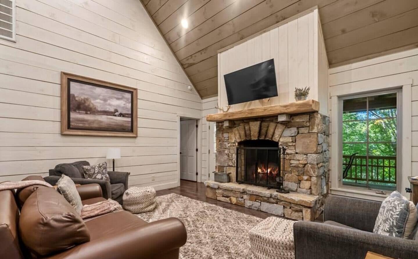 A cozy living room with a stone fireplace, wooden walls, and a large window overlooking greenery.