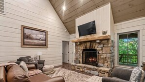 Un salon chaleureux avec une cheminée en pierre, des murs en bois et une grande fenêtre donnant sur un paysage verdoyant.