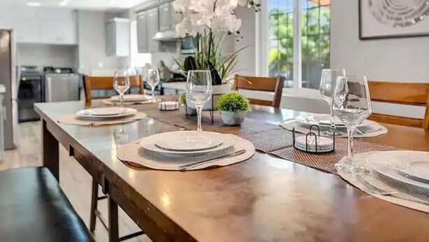 Een moderne eetruimte met een lange tafel gedekt voor twee personen, omringd door stoelen, en een keuken op de achtergrond.