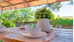 Een buitenterras met een houten tafel gedekt voor twee personen, een witte vaas met groene begroeiing, en een uitzicht op groen in de achtergrond.