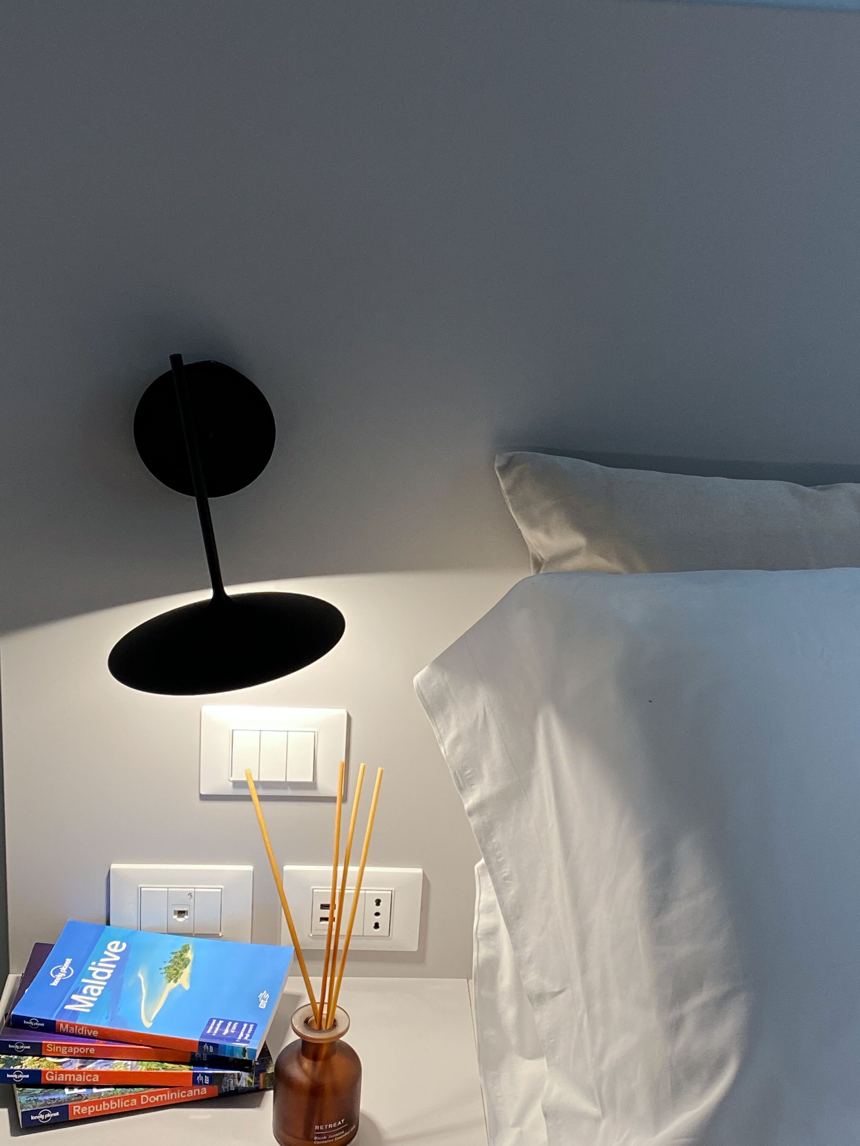A neatly made bed with white sheets, a bedside table with a stack of travel books, a black wall-mounted lamp, and a white power outlet.