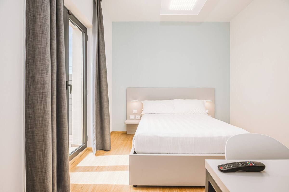 Une chambre d’hôtel avec un lit, un bureau, une télécommande et une fenêtre avec des rideaux.