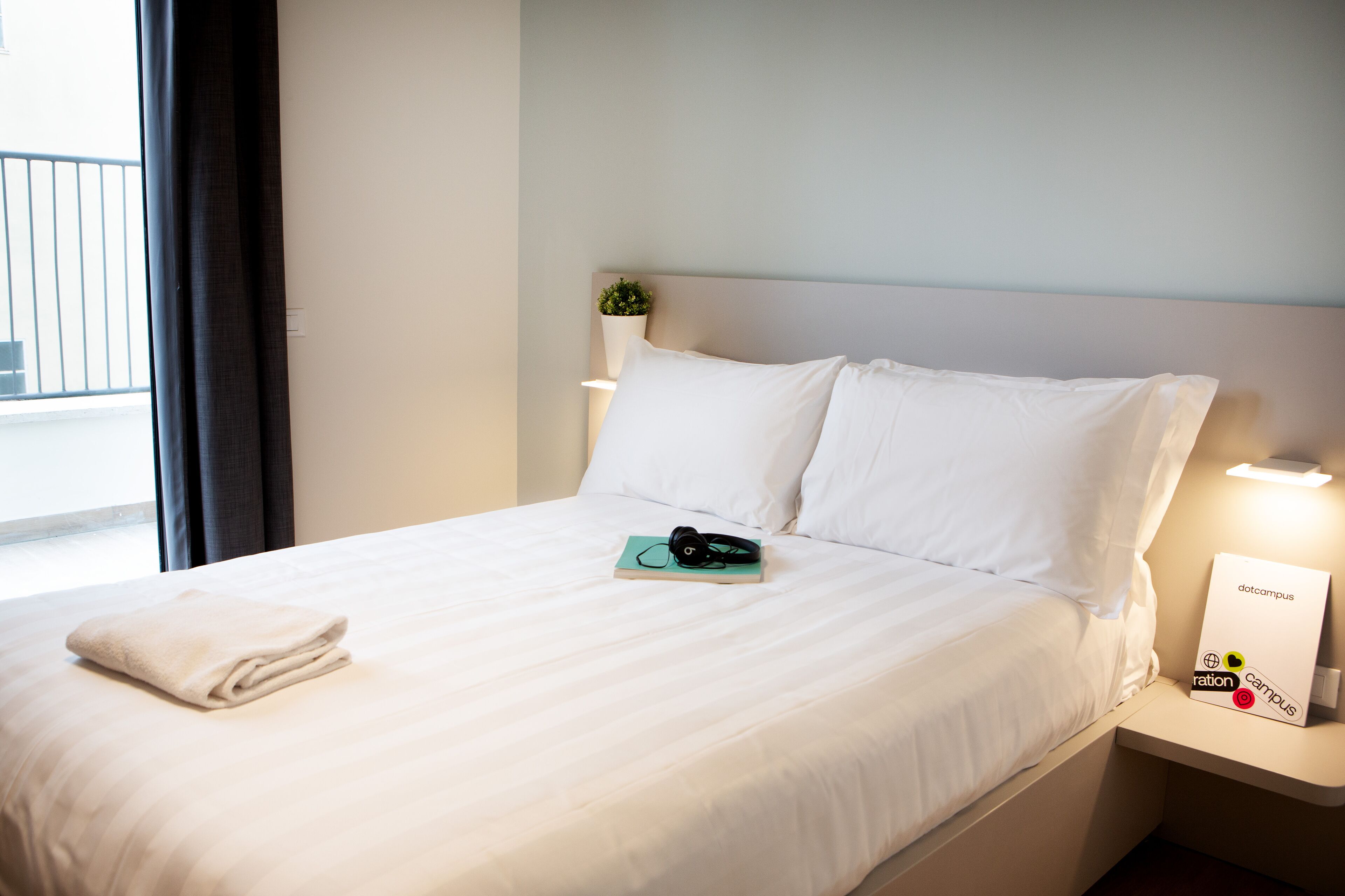 A neatly made bed with a folded towel, a pair of headphones, and a sign on the nightstand.