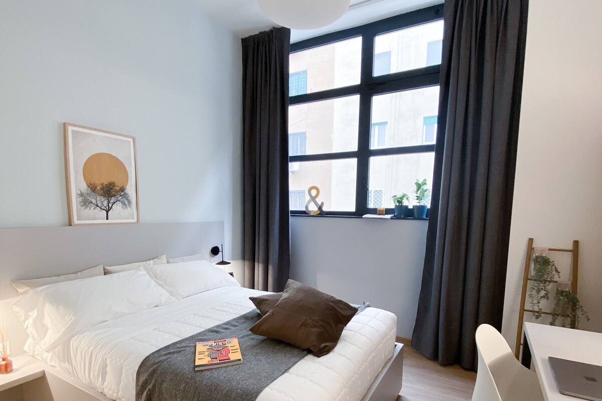 Une chambre à coucher moderne avec un lit, une table de chevet et un bureau avec une chaise. Il y a une fenêtre avec des rideaux, un luminaire suspendu et un tableau encadré au mur.