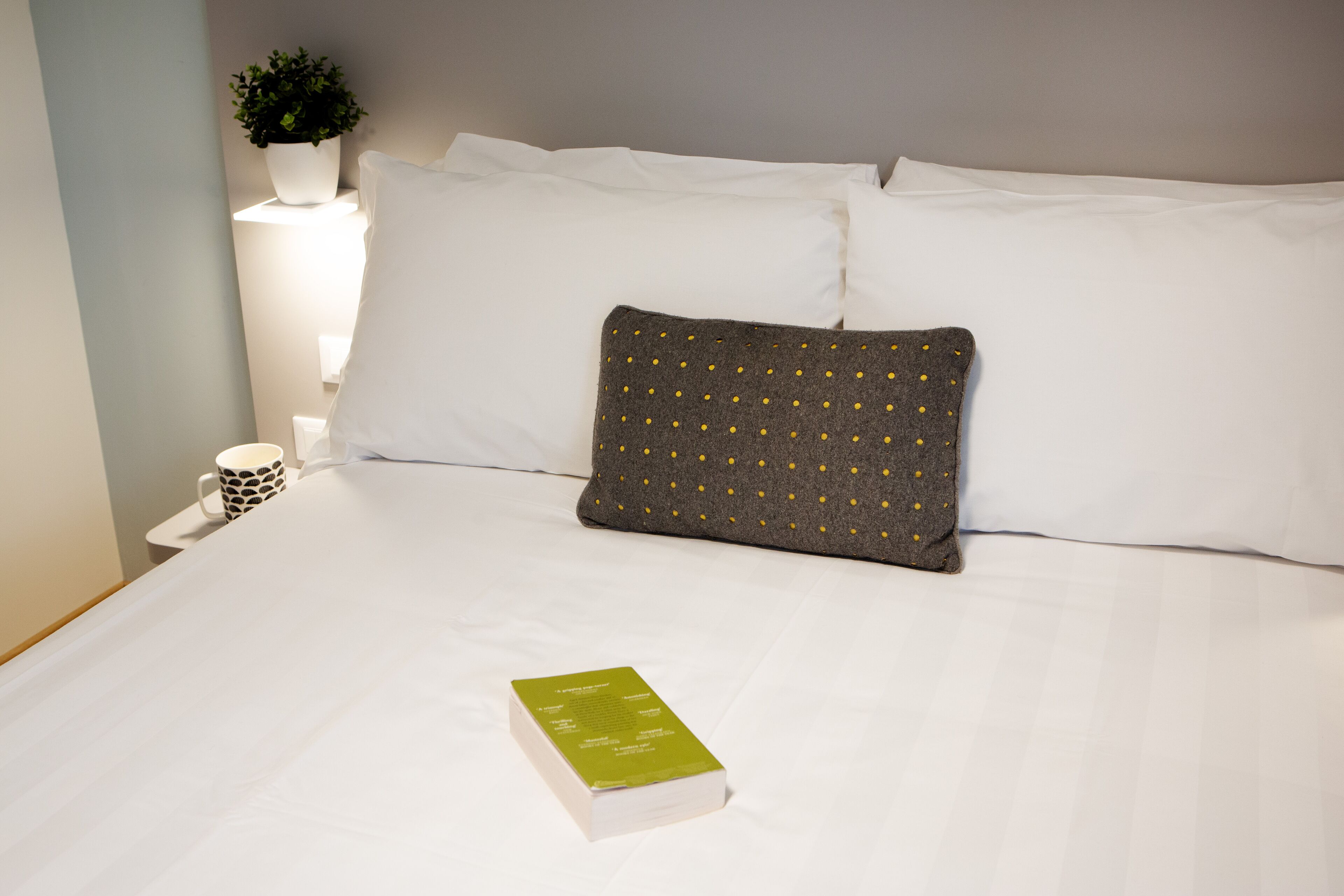A neatly made bed with white linens, a single green book, and a gray pillow with yellow dots.