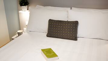 A neatly made bed with white linens, a single green book, and a gray pillow with yellow dots.