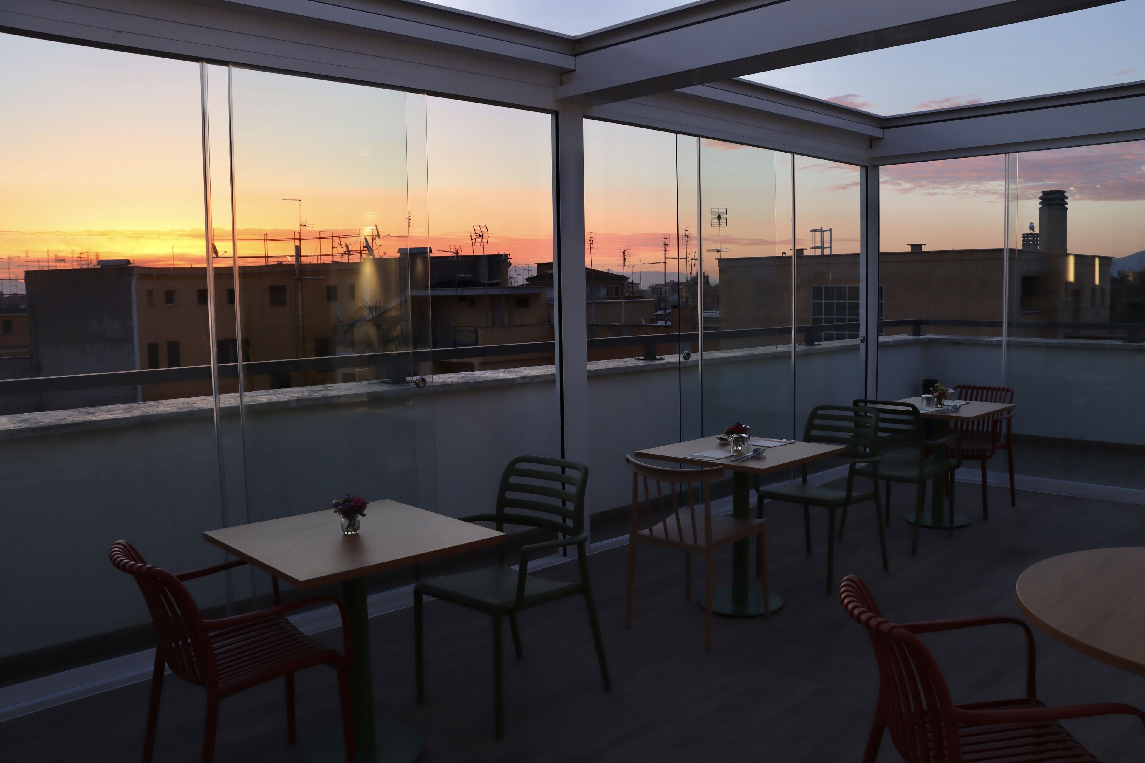 A modern rooftop terrace with glass walls, offering views of the city skyline at sunset.