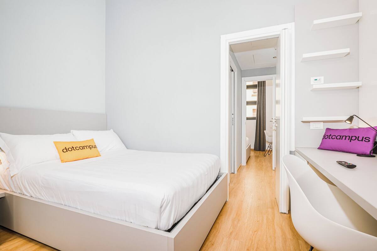 Une chambre d’hôtel moderne avec un lit blanc, un bureau agrémenté d’un coussin violet et une porte visible donnant sur une autre pièce.