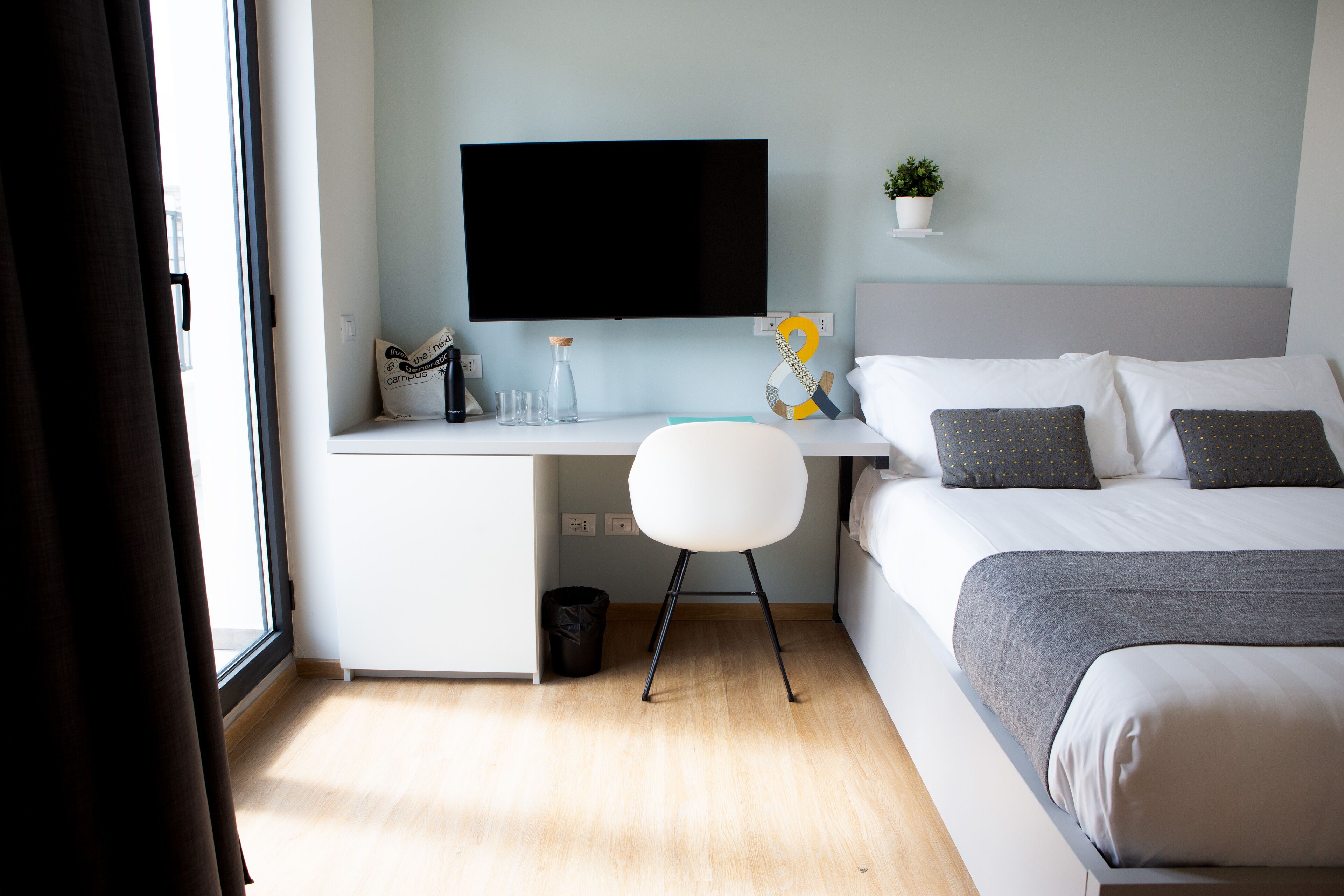 Habitación de hotel con cama, escritorio con silla, televisor, planta pequeña y ventana.