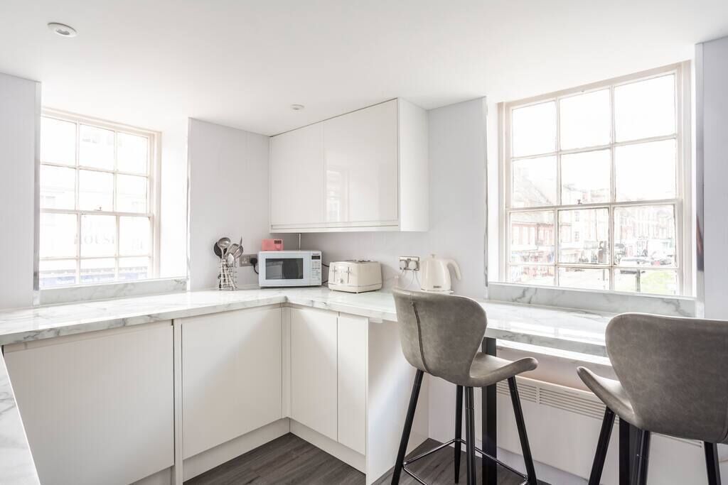 Eine moderne Küche mit weißen Schränken, einer Mikrowelle, einem Toaster und einer Kaffeemaschine auf der Arbeitsplatte. Zwei Barhocker und große Fenster sorgen für natürliches Licht.