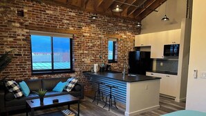 A modern kitchen with a brick wall, a dining area with a sofa and coffee table, and a large window. - Arch Building Lofts - Unit 6 (Redmond)