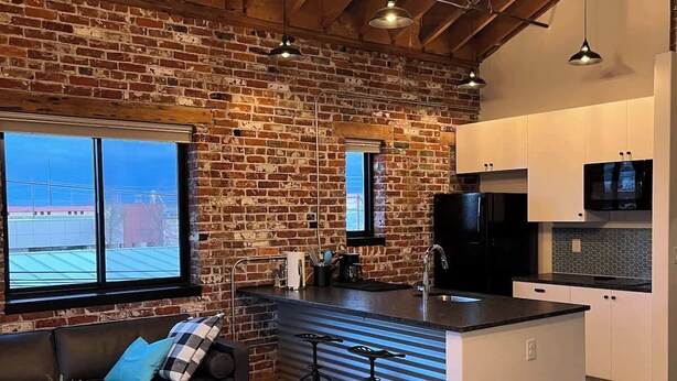 A cozy living area with a brick wall, wooden ceiling, and a large window. There is a sofa, a coffee table, and a kitchen with white cabinets and a black countertop. - Arch Building Lofts - Unit 6 (Redmond)