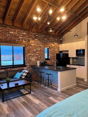Una sala de estar acogedora con pared de ladrillo, techo de madera y un ventanal grande. Se observa un sofá, una mesa de centro y una cocina con armarios blancos y encimera negra.