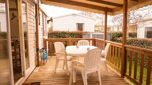 A covered patio with a wooden deck, white plastic chairs, and a round table. - FAMILY MOBILHOME 6/8 pers COMFORTABLE AND AIR CONDITIONED (Veuzain-sur-Loire)