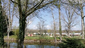 A serene lakeside setting with trees, a pond, and a row of white buildings in the background. - FAMILY MOBILHOME 6/8 pers COMFORTABLE AND AIR CONDITIONED (Veuzain-sur-Loire)