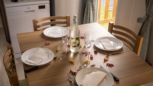 A dining table set with plates, cutlery, a bottle, and glasses, with a view of a kitchen and a window in the background. - FAMILY MOBILHOME 6/8 pers COMFORTABLE AND AIR CONDITIONED (Veuzain-sur-Loire)