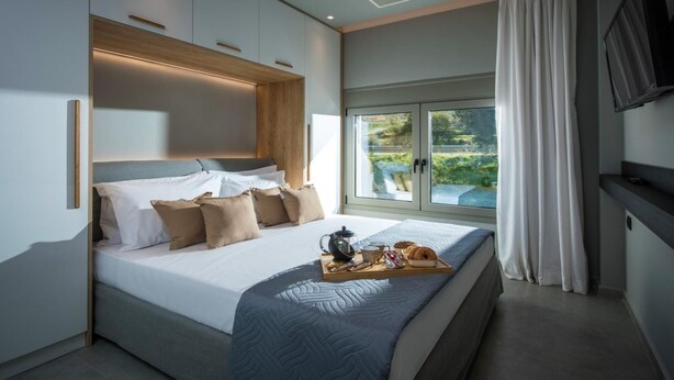 A modern bedroom with a large bed, a bedside table with a tray of breakfast, and a TV mounted on the wall. - Malia Palace by Luxury Living Apartments (Hersonissos)