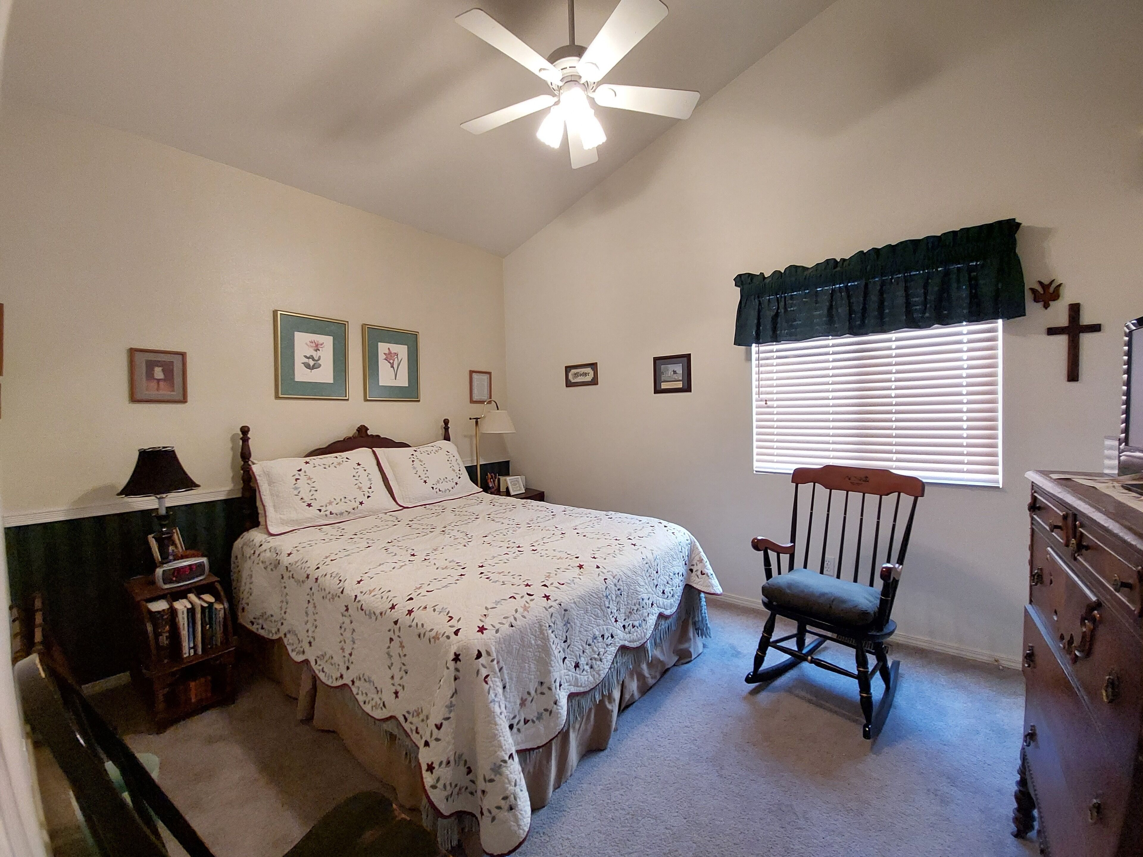 A bedroom with a bed, a rocking chair, a dresser, a nightstand, a lamp, a ceiling fan, and a window with blinds.