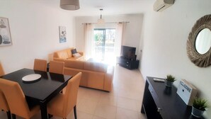 A modern living room with a sofa, a dining table with chairs, a TV, and a small desk with a sign. - Apartment Buganvilia Porto de Mós (Lagos)