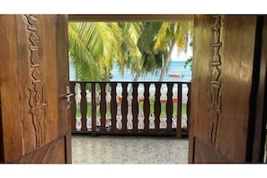 A wooden door with intricate carvings, leading to a balcony with a view of palm trees and the sea.